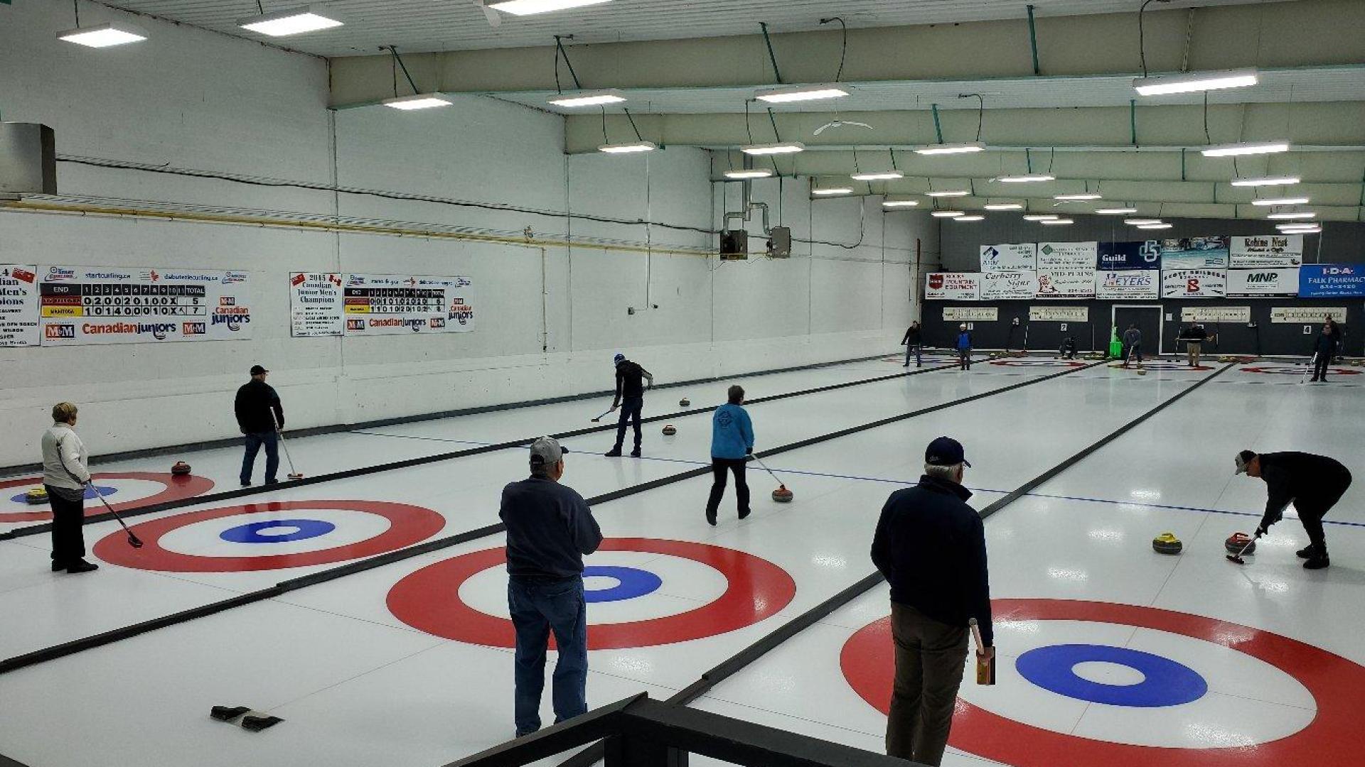 A group of people in warm clothing on the ice at a curling rink. All lanes are occupied by the people playing. 