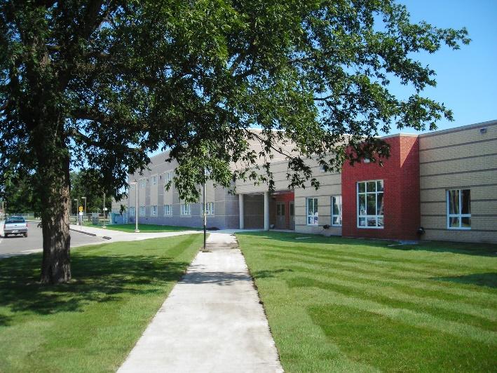 The facade of Carberry Collegiate, a tan and red building, viewed from a path, with a large tree in the foreground. 