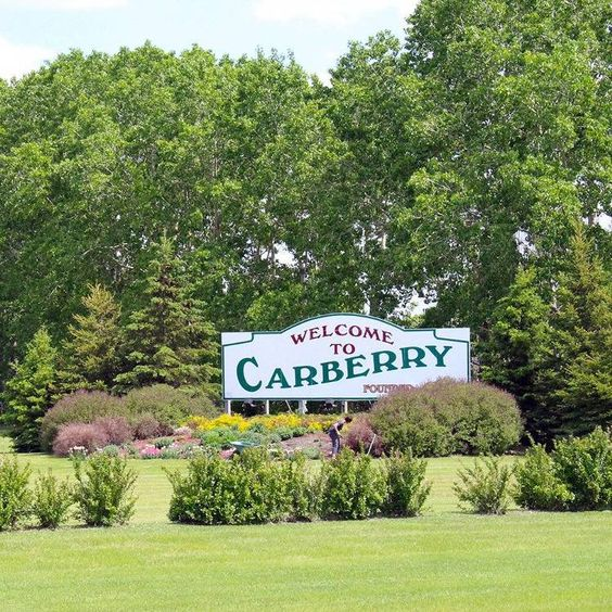 A greenscape with shrubs and trees, the focus is a sign reading Welcome to Carberry.