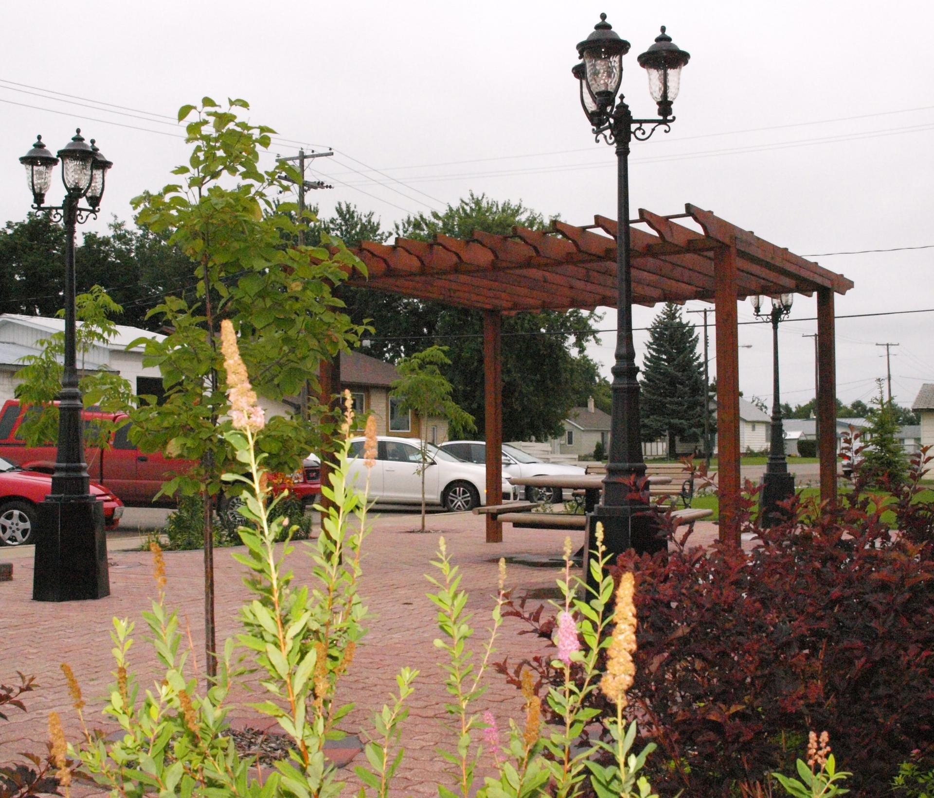 A town square paved with bricks, with a metal pergola and old-fashioned streetlamps and flowers in the foreground.
