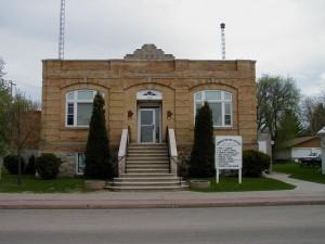 Carberry old town hall, a short, square brick building with a staircase leading to the front door and a small sign out front. 